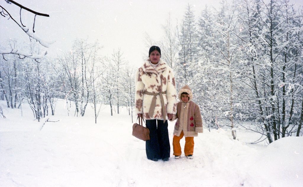 Indisk mor i pelskåpe med et lite barn i vinterdress i snødekt norsk skog, 1970-tallet